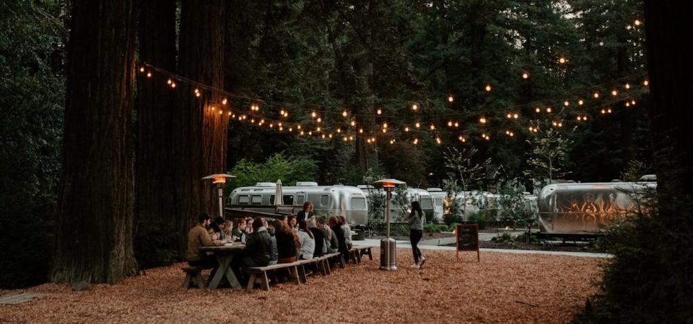 Group dining at a picnic table in a redwood grove at twilight, café string lights overhead, and polished Airstream trailers parked beyond