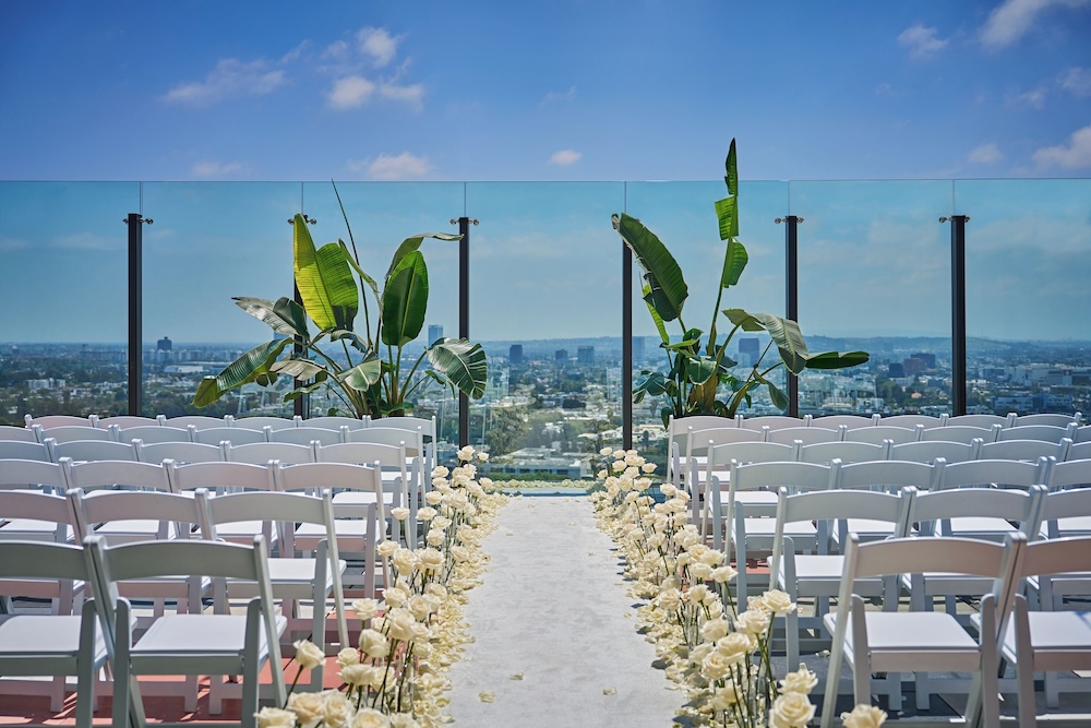 Rooftop ceremony with white folding chairs flanking a petal-strewn aisle, bird-of-paradise framing the altar, LA cityscape beyond a glass railing