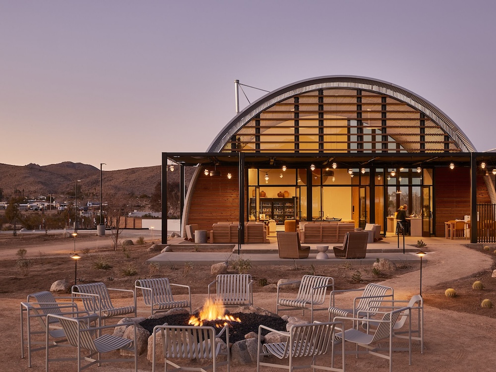 Arched Quonset-style desert clubhouse glowing at dusk, a ring of steel slatted chairs around a stone fire pit, and Joshua Tree mountains behind