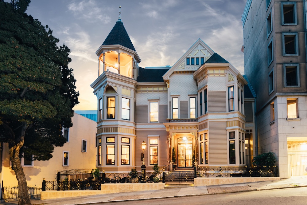 Cream Victorian mansion at twilight with a conical-roofed corner turret, peaked gable, glowing bay windows, and an iron-fenced front entrance