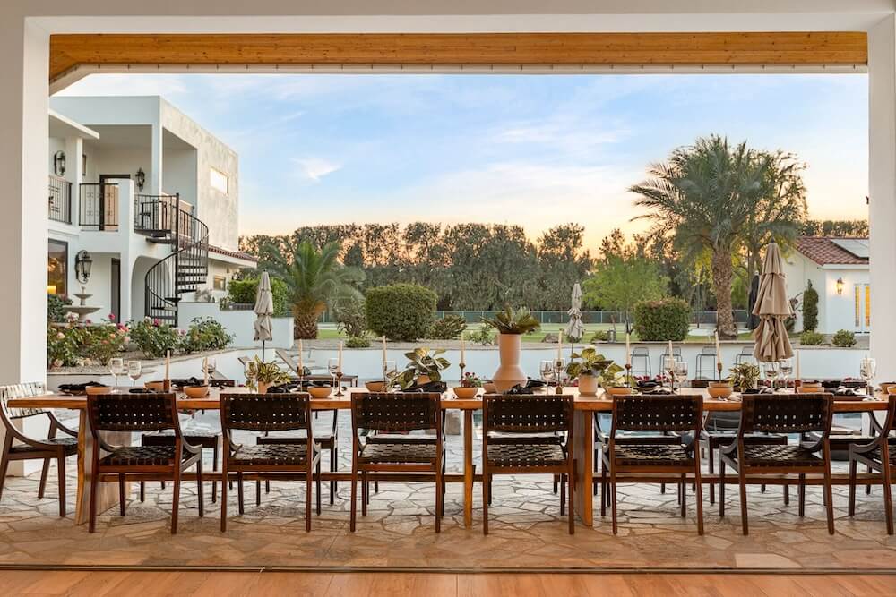 Long wood reception table with woven leather chairs and candles under an open portico, view of a Mediterranean villa, pool and palms at sunset