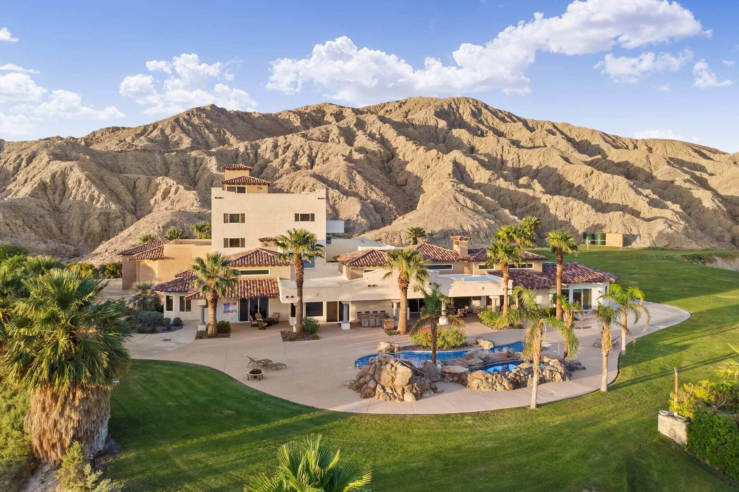 Aerial view of a Mediterranean desert estate with terracotta roofs and palms around a curved pool, eroded mountains rising directly behind