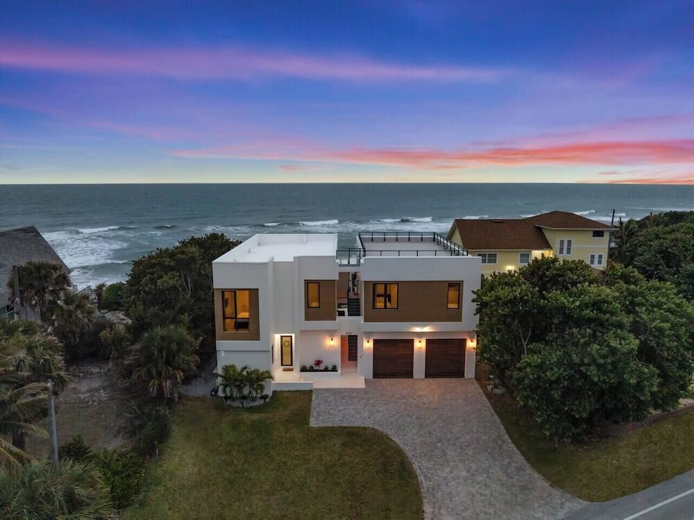 Aerial twilight view of a modern white-stucco beachfront home with rooftop deck, pink-and-purple sunset sky, Atlantic surf in the distance