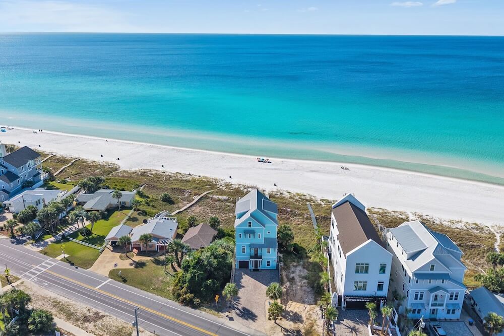 Aerial Panama City Beach view of a pale blue three-story stilted beachfront home, white sand dunes in front, turquoise Gulf water beyond