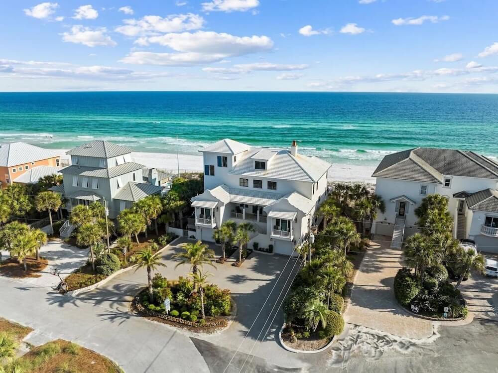 Aerial daytime view of a three-story white beachfront estate with multi-level porches, circular paver driveway, palms, turquoise Gulf beyond