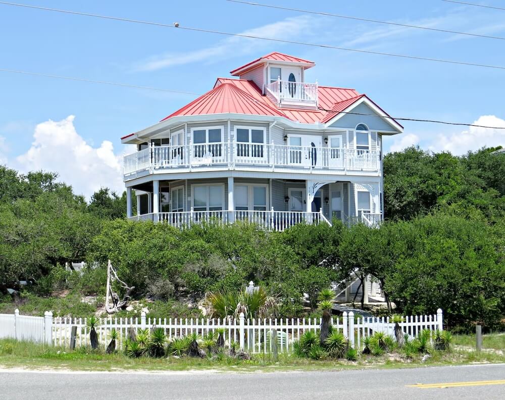 Victorian-style coastal home with white siding, a two-tier wraparound porch, red metal roof, rooftop cupola, white picket fence and yucca plants