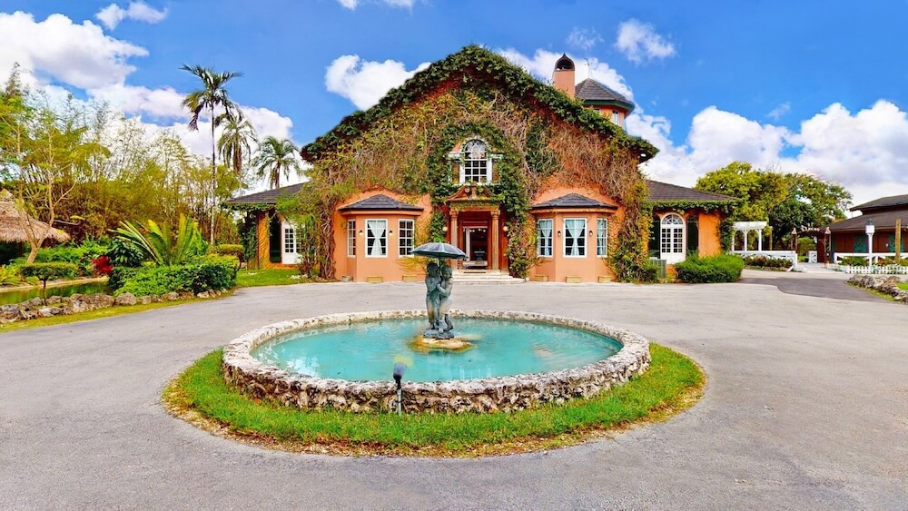 Ivy-covered terracotta Mediterranean estate with a circular driveway and sculpted stone fountain at center, tropical foliage and palms framing