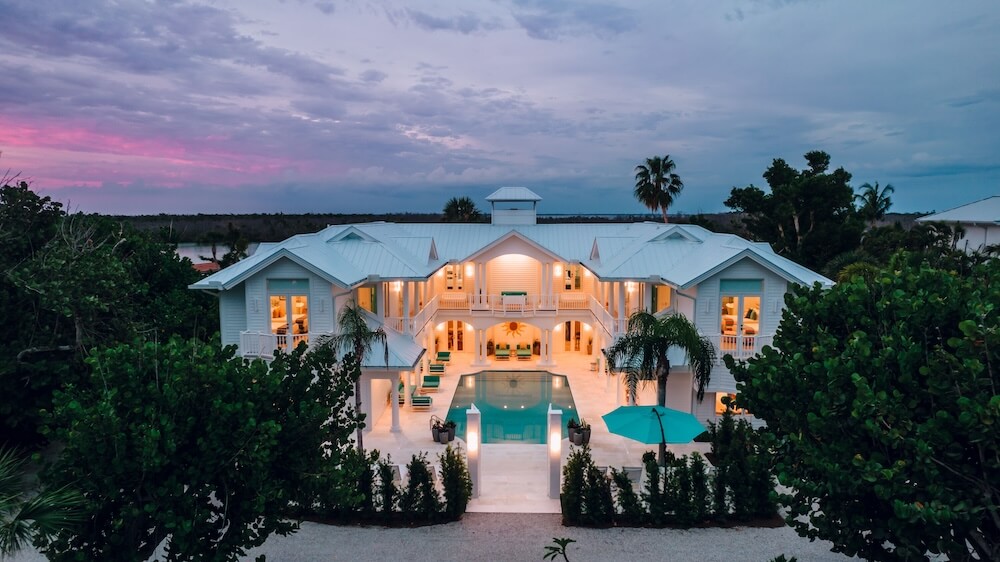 Grand symmetrical white coastal mansion at twilight, opposing wings flanking a sunken rectangular pool, sea grape hedges and palms, pink sunset