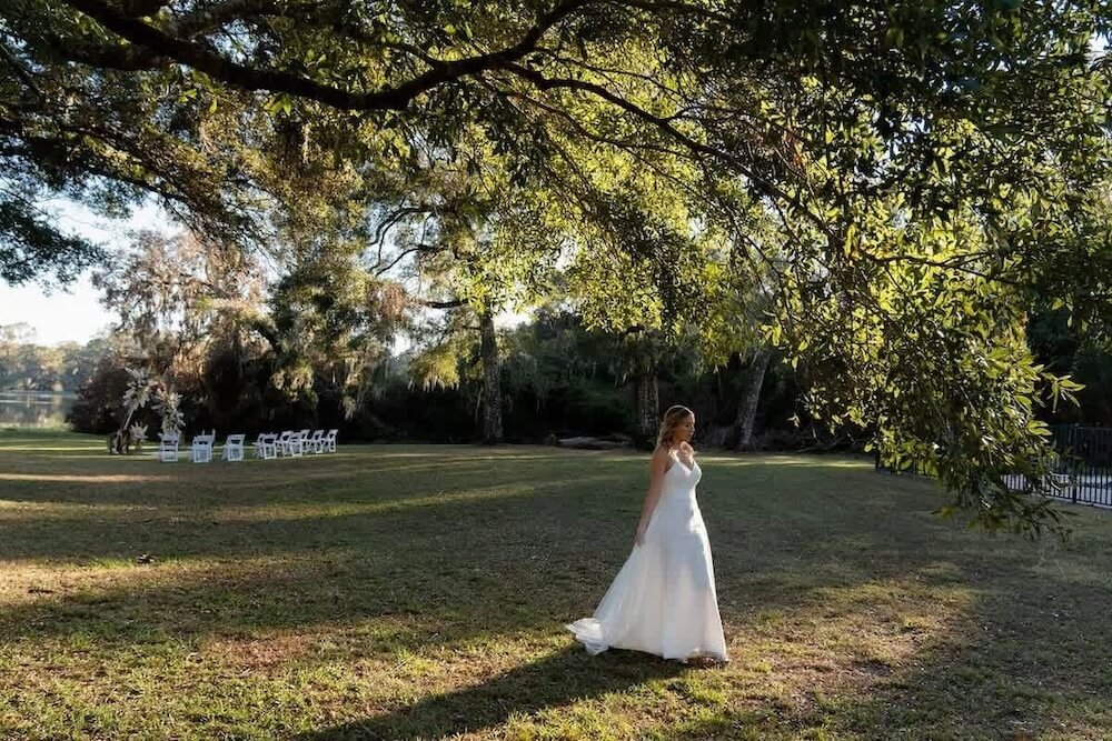 Person in a white wedding gown standing alone under a massive Spanish-moss-draped live oak, ceremony chairs set up across the lawn toward a lake