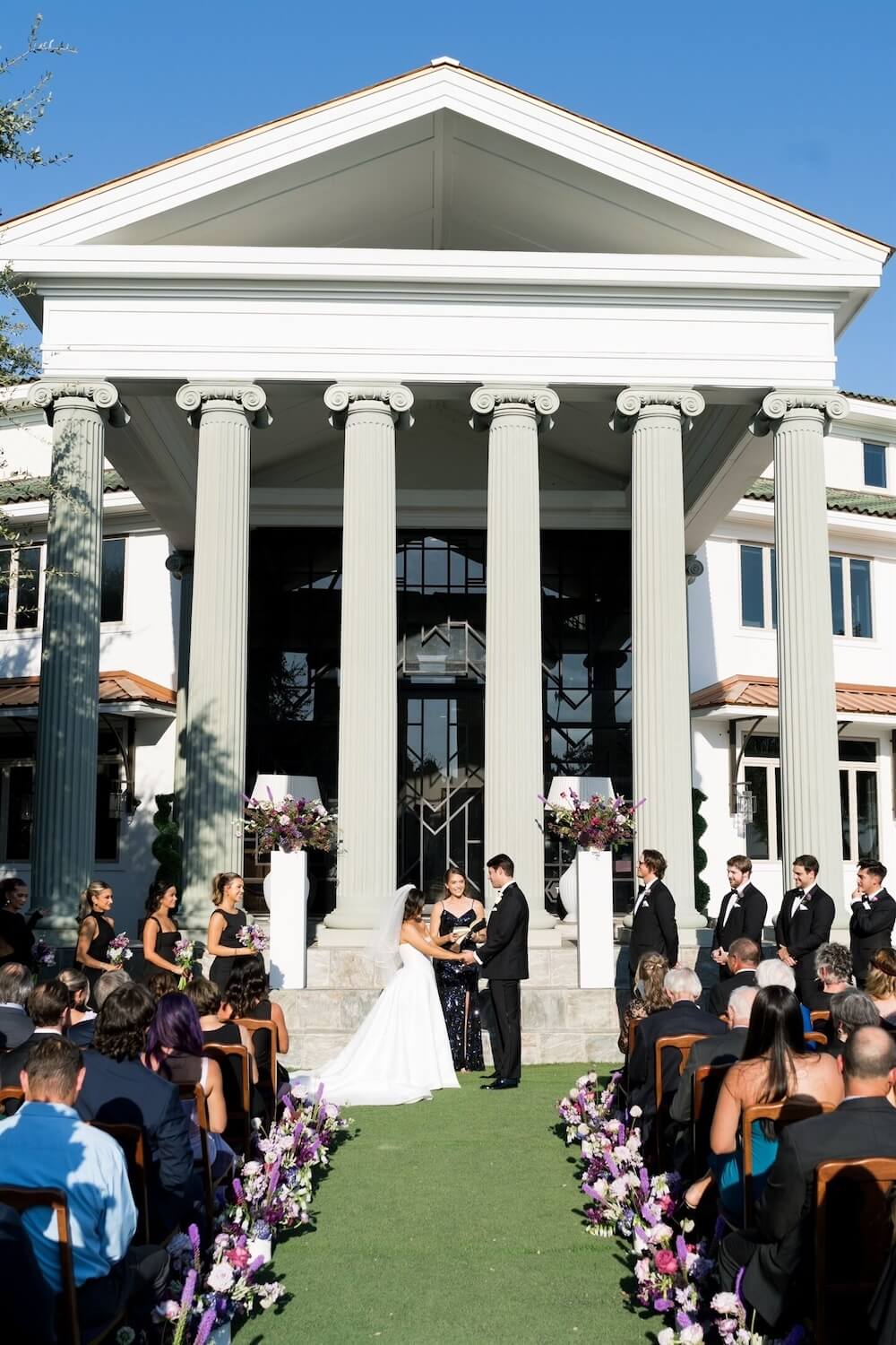 Outdoor ceremony before a Greek-Revival VRBO wedding venue with Ionic columns, couple at the altar on a lawn aisle lined with purple florals