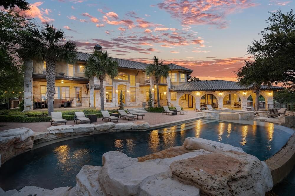 Mediterranean limestone VRBO wedding villa at sunset, peach-streaked sky, palms flanking a free-form pool with stone-edged decking