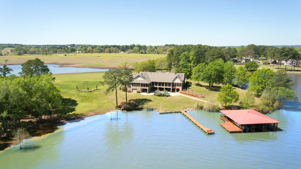 Aerial of a brick lakefront VRBO wedding venue, mature trees on the lawn, a long wooden dock to a red-roofed covered boathouse