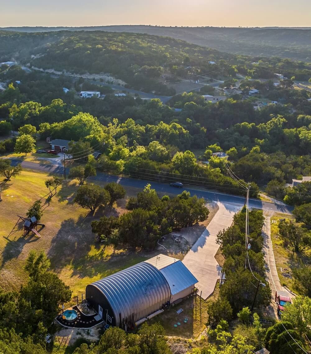 Aerial of an arched metal-roof Hill Country VRBO wedding venue, oaks and rolling hills around with a small pool out front