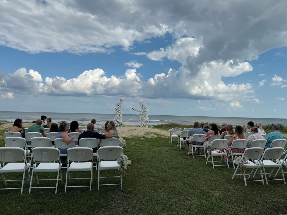 Beachfront ceremony at a Gulf Coast VRBO wedding venue, guests in two banks of folding chairs facing a white-floral arch, puffy clouds beyond
