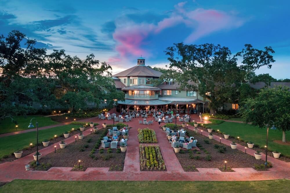 Twilight aerial of an Alabama bayfront small wedding venue, cupola-topped rotunda, formal parterre garden, lit lampposts, pink wisp of sunset cloud above