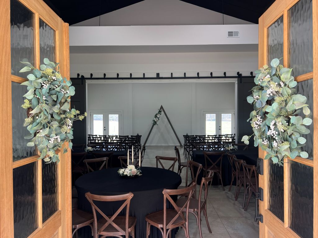 Eucalyptus-wreathed wood doors open into a modern Alabama small wedding venue, black round tables and cross-back chairs, triangular wood ceremony arch