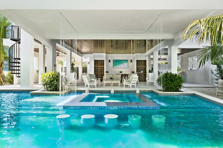 Turquoise pool with submerged barstools at a swim-up bar under a covered loggia, palms and a black spiral staircase at the edge of a luxury home