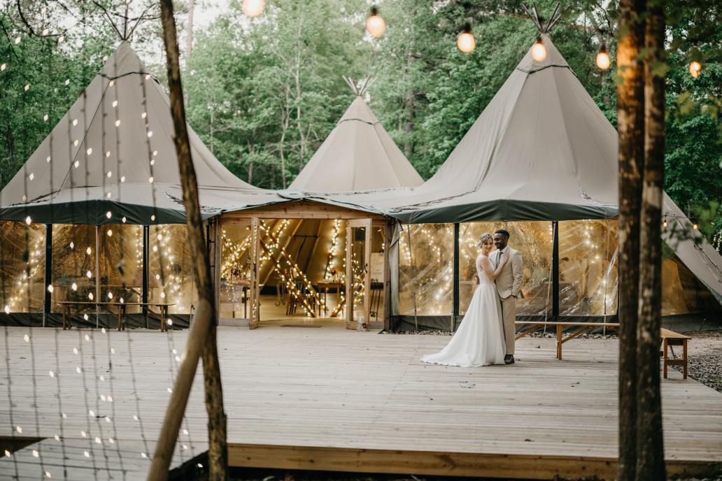 Couple in wedding attire on a wood deck in front of a cluster of Nordic-style canvas tipi tents glowing with fairy lights, deep forest behind