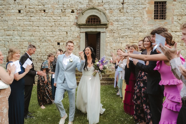 Couple walking laughing through a parted aisle of cheering guests after their small wedding in Italy, stone castle façade with arched doorway behind