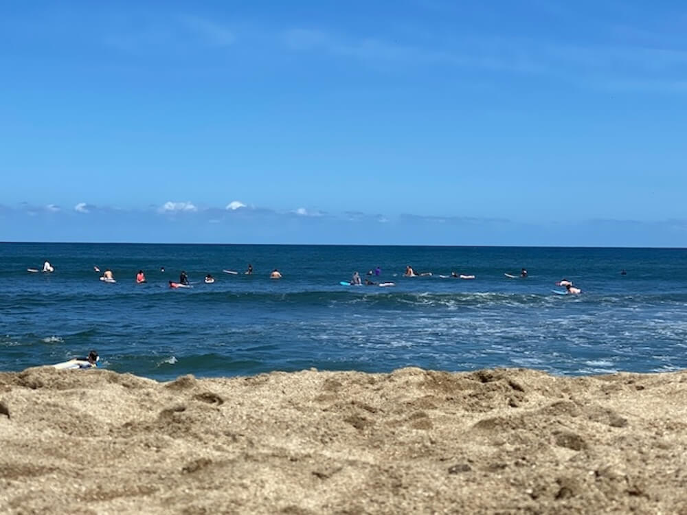 Wide view from the sand of surfers lined up in the break at the main Sayulita beach, sandy foreground and calm blue Pacific behind