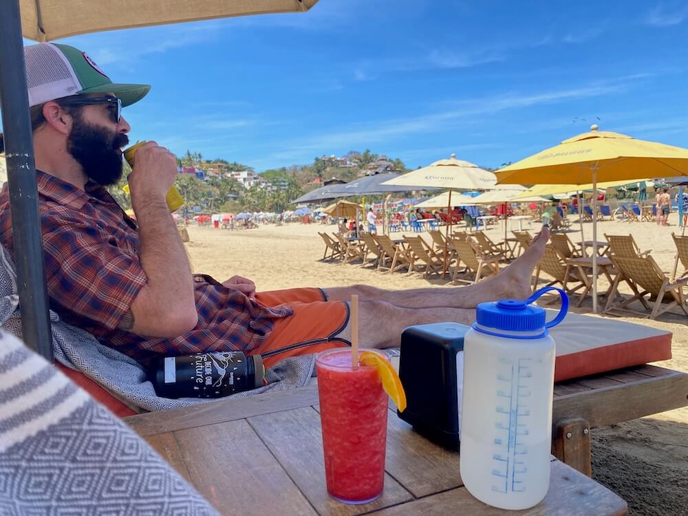 Person reclining in a Sayulita beach-club lounge chair under shade with a frozen drink and water bottle, rows of yellow umbrellas behind
