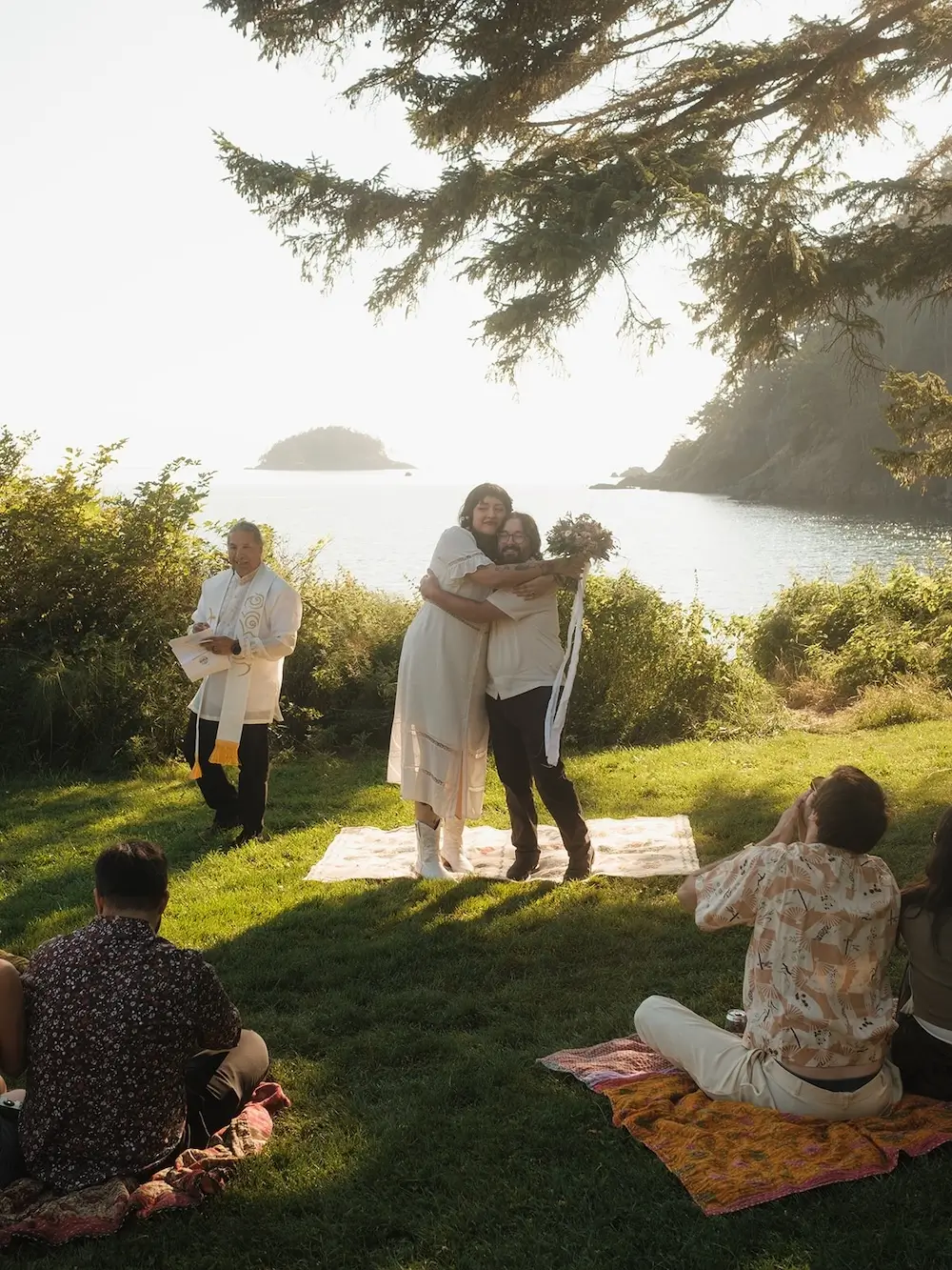 Intimate outdoor wedding ceremony on a grassy coastal overlook with guests seated on blankets as the couple embraces beside the officiant overlooking the water.