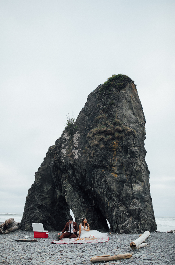 Couple shares a picnic on a sandy beach during their elopement, with rocky coastline behind them