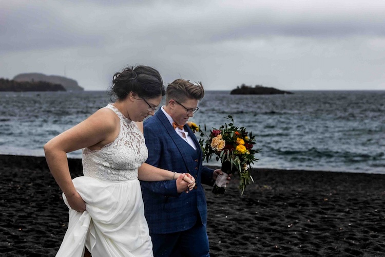 Couple walks together along a rocky coastline after their small beach wedding, water in the background