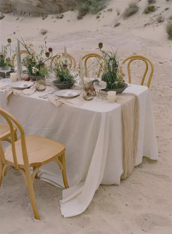 Long table set with linens and tableware for an intimate beach wedding reception, sand and dune grass beyond