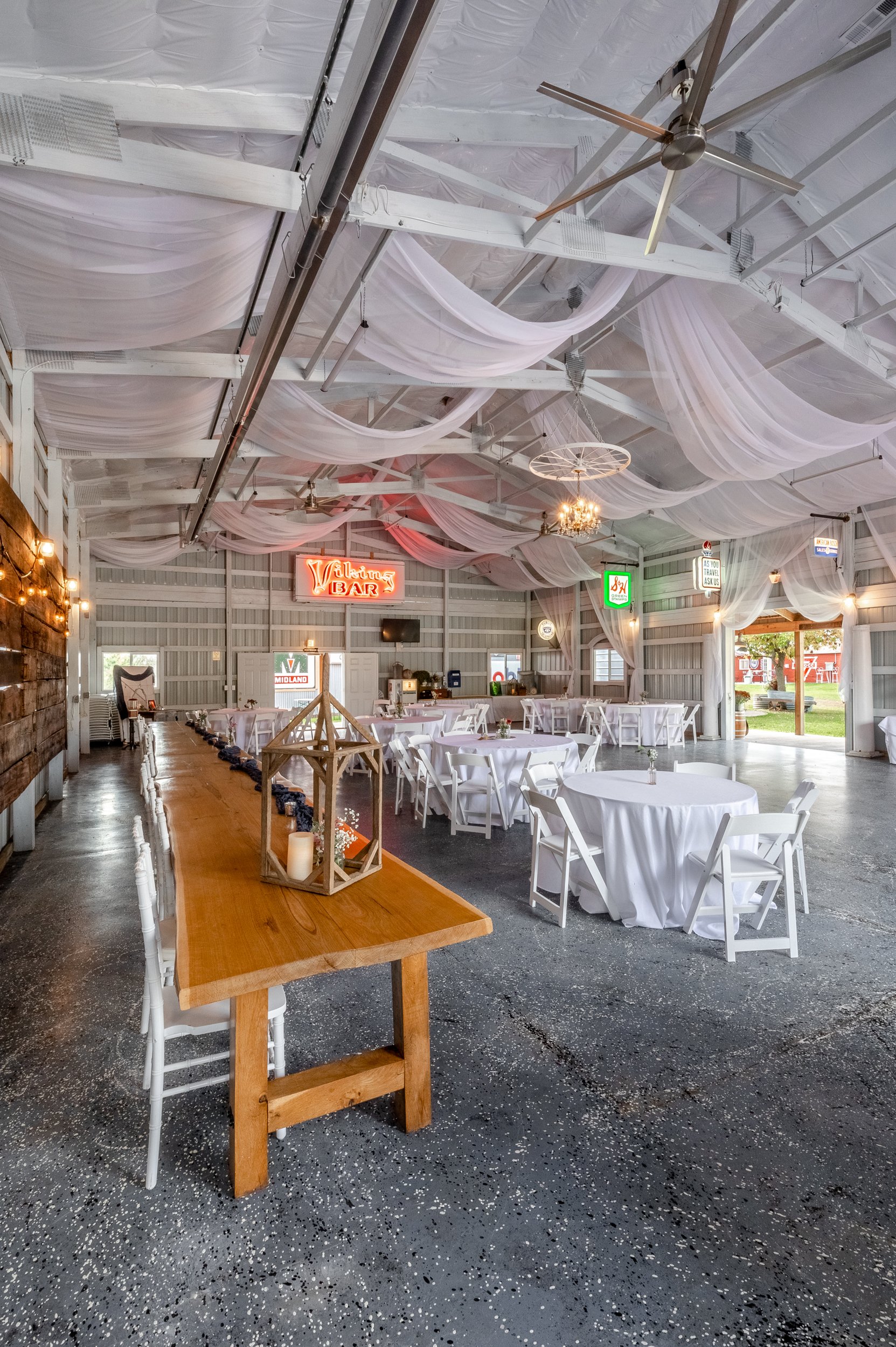 Long farm table with lantern centerpieces in an Adler Ranch reception barn, white drapery swagged from rafters, vintage 'BAR' neon sign on the back wall