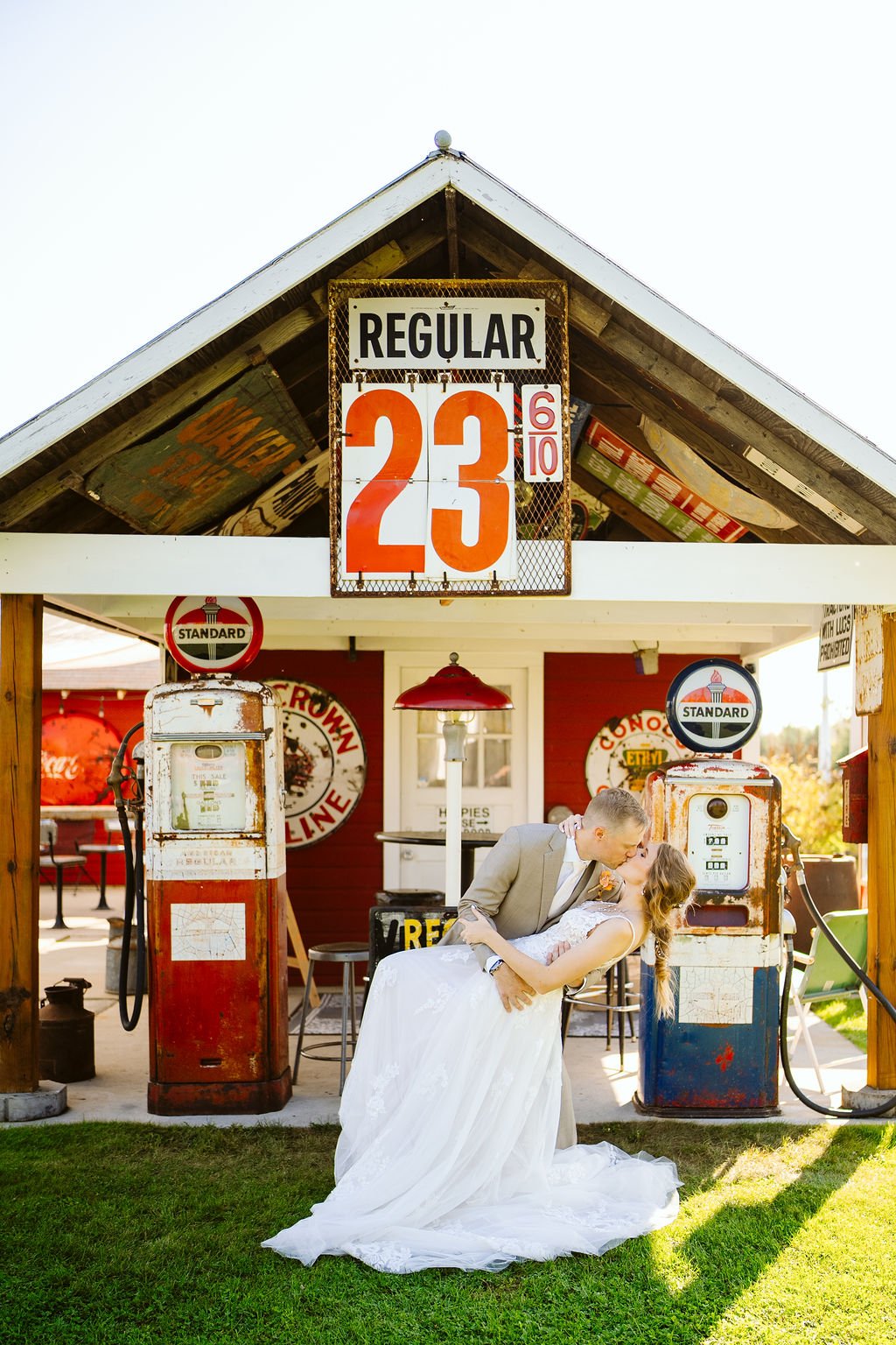 Couple in a dip kiss at an Alexandria Minnesota small wedding venue, vintage red filling station behind them with antique gas pumps and a 'REGULAR 23' sign