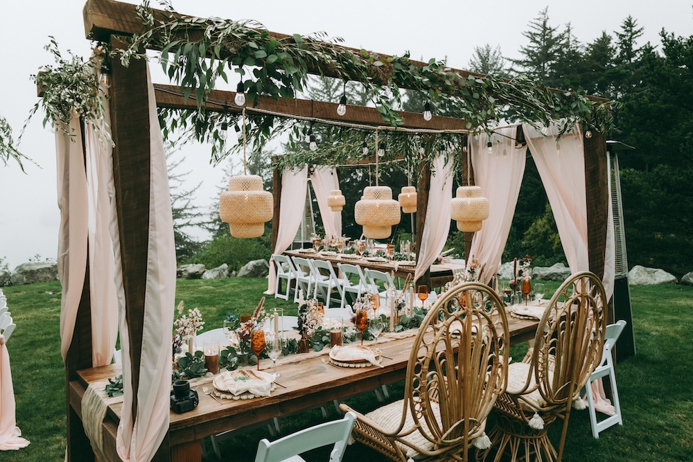 Boho coastal reception, long wood table under a pergola with eucalyptus and cream curtains, bamboo pendant lanterns, peacock rattan head chairs