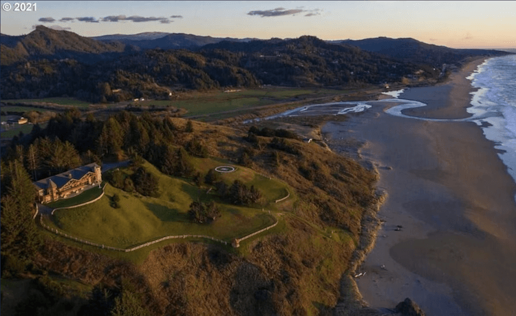 Aerial of an Oregon Coast VRBO wedding venue at golden hour, log home on a bluff with meadow lawn, helipad, beach curving into the distance