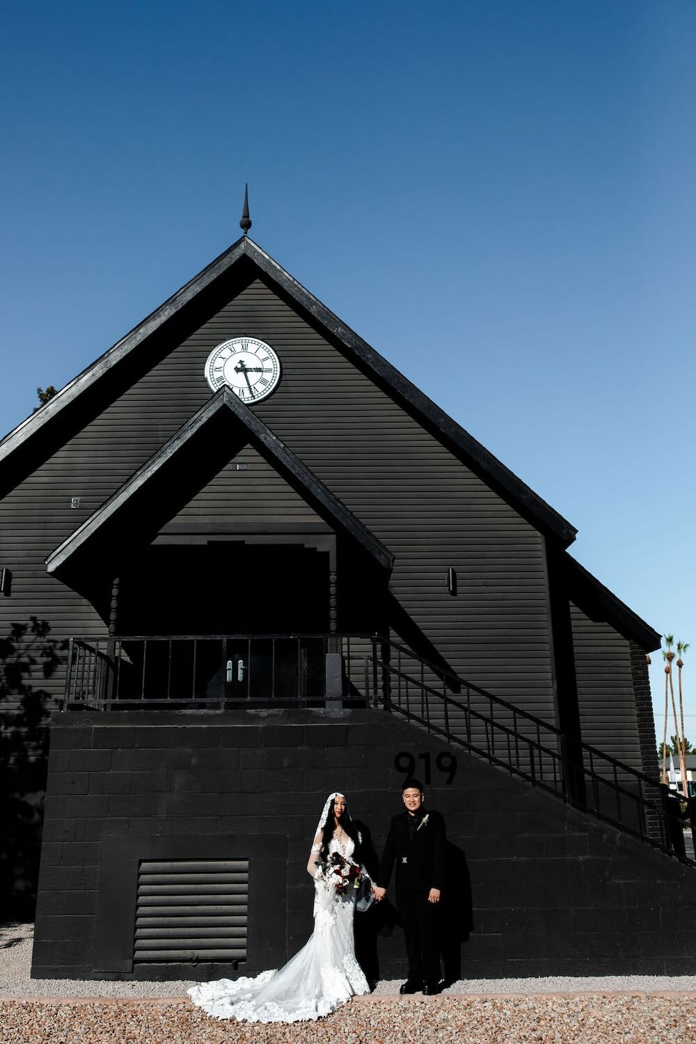 A couple stands dressed in wedding attire in front of a black wedding chapel