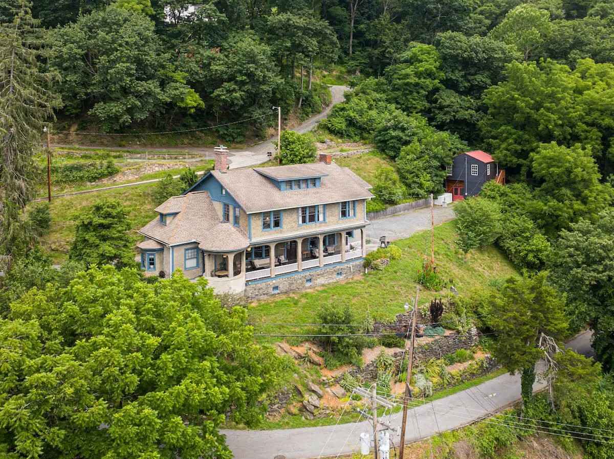 Aerial of a Shingle-style Asheville-area VRBO wedding rental with blue trim and wraparound porch above the French Broad River, terraced gardens below