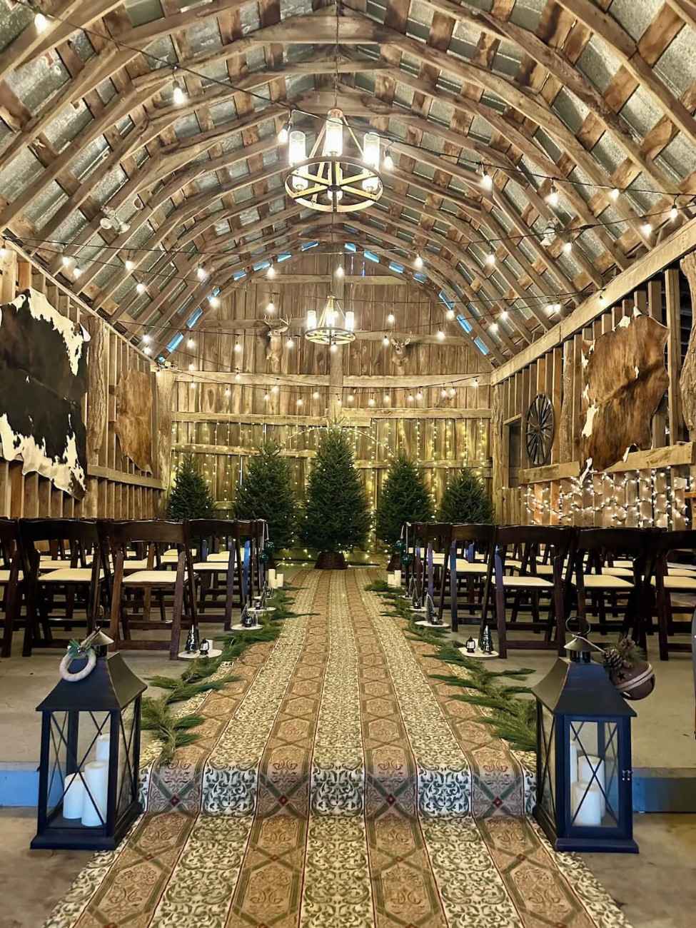 Historic NC barn interior with arched timber ceiling and wagon-wheel chandeliers, damask aisle runner between dark wood folding chairs