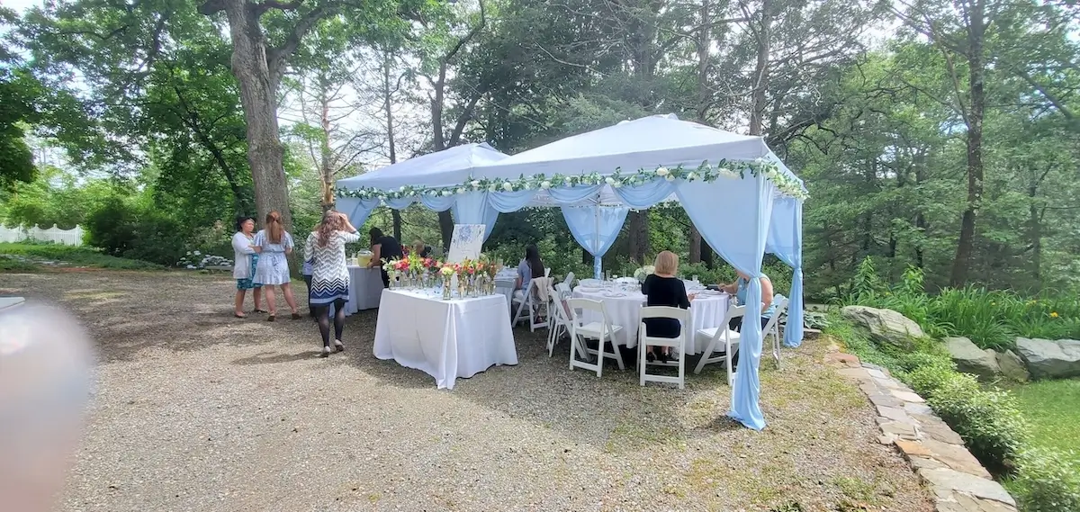 Pale-blue draped wedding canopies with green-and-white floral garlands over a gravel terrace, white-linen tables underneath, stone wall bordering forest