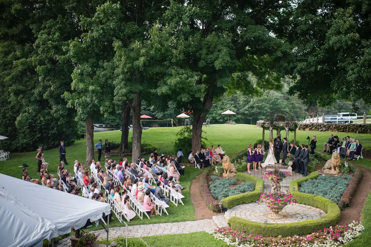 Massachusetts VRBO wedding venue garden ceremony — couple at a boxwood-hedge altar with tiered fountain and stone lions, guests on white folding chairs
