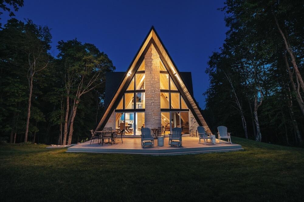 Glowing A-frame VRBO rental at dusk, timber-cathedral ceiling through the glass front, central stone chimney, Adirondack chairs on the curved patio