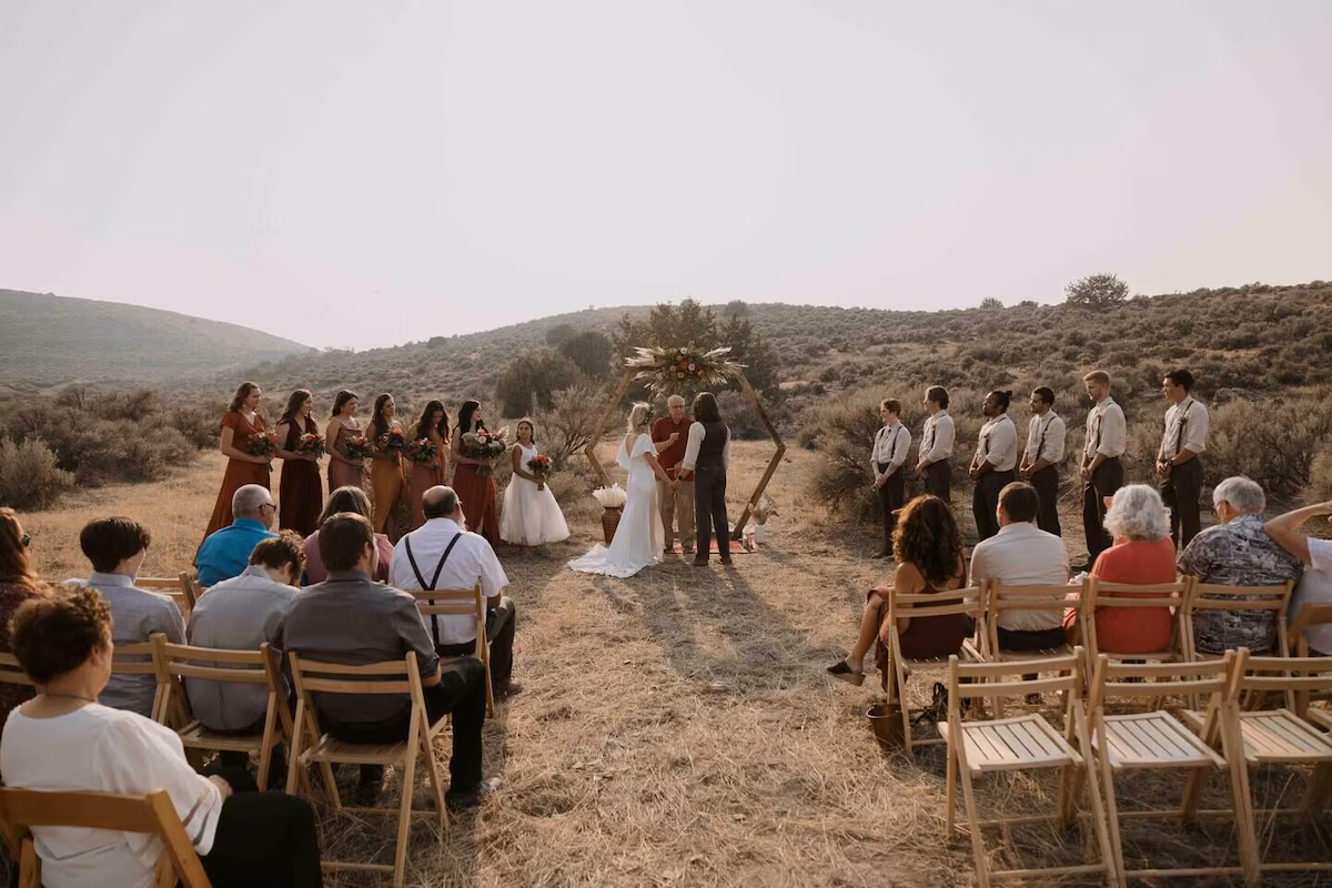 Sagebrush ceremony — couple at a hexagonal wood arch with pampas and burnt-orange florals, wedding party in rust, dried sage hills under golden sky