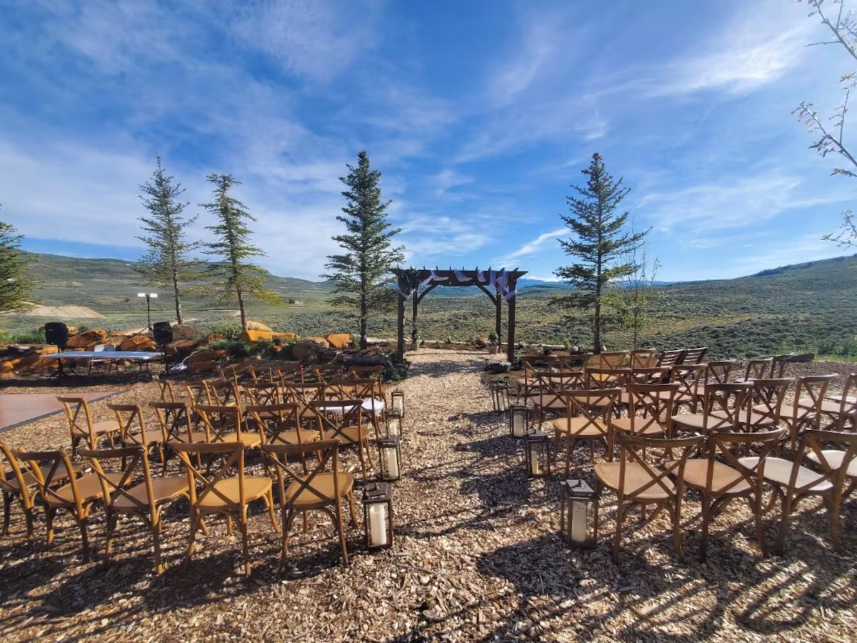 Mountaintop ceremony — cross-back chairs facing a dark wood arbor with greenery, hurricane lanterns, evergreens framing a Wasatch valley view