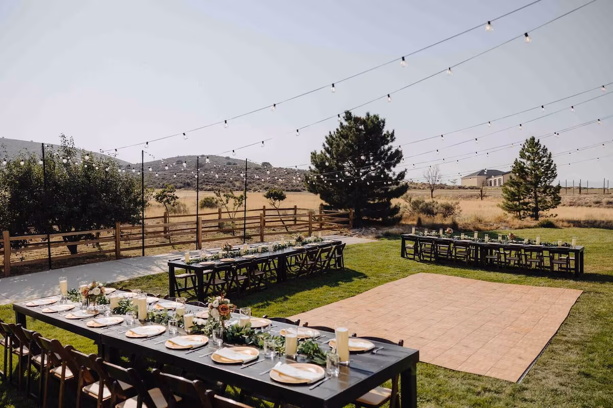 Outdoor reception — three long dark farm tables in a U with gold chargers and greenery, wood dance floor, bistro lights, sagebrush field beyond