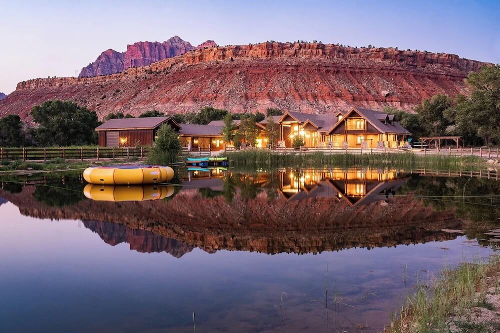 Dark timber Zion VRBO wedding lodges reflected in a glassy pond at dusk, yellow water trampoline, red rock cliffs catching sunset light