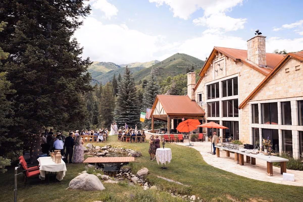 Wedding ceremony on a lawn behind a stone Park City VRBO wedding lodge, footbridge over a creek, red patio umbrellas, Wasatch peaks rising behind