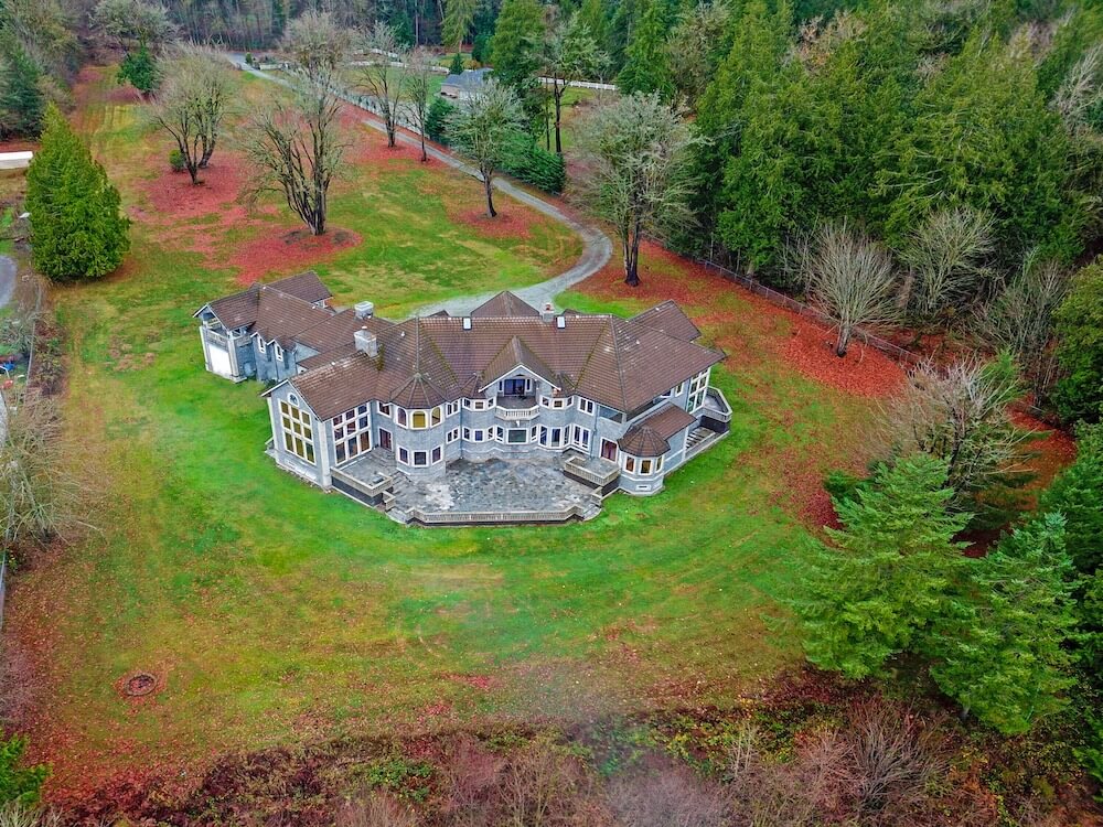 Aerial of a gray-stone Pacific Northwest castle with gables and bay windows, green lawns and rust-orange leaves at the edges, evergreens framing