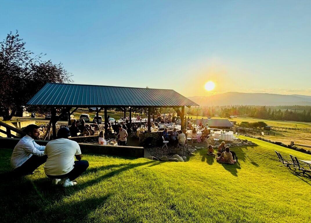 Wedding reception under a covered metal-roof pavilion at sunset, guests dining, sun setting over the Eastern Washington valley and distant ridges