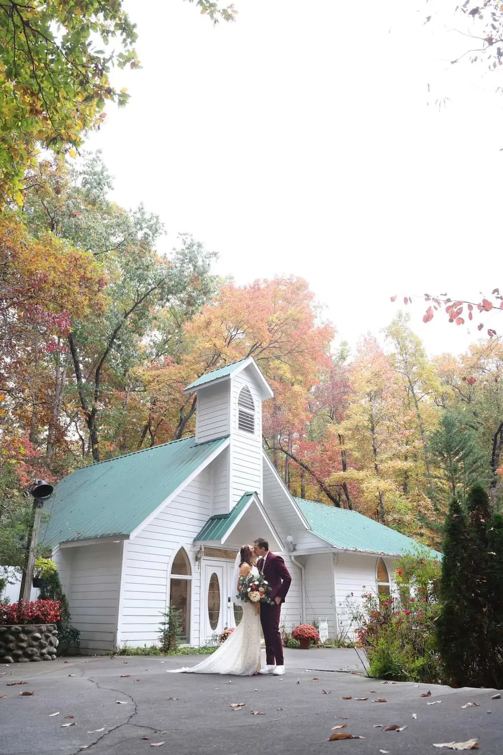 Couple kissing in front of the white chapel at Chapel at the Park micro wedding venue surrounded by fall foliage in Gatlinburg, Tennessee.