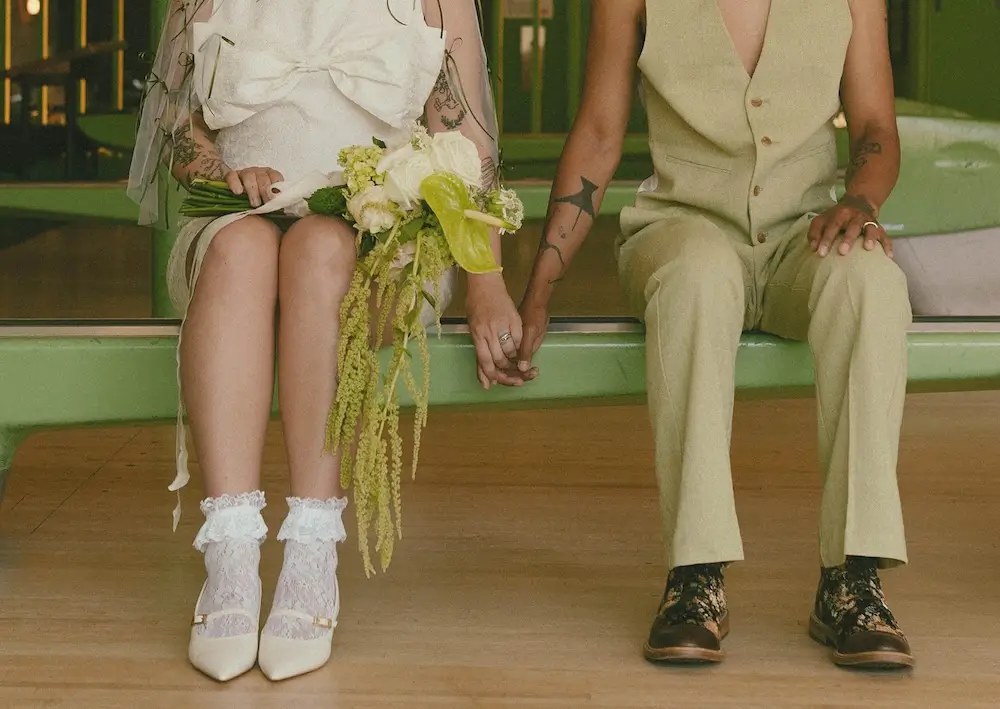 Close-up of a couple seated on a bowling alley ball return, holding hands in modern wedding attire with a green-and-white bouquet and vintage-inspired lace socks and loafers.