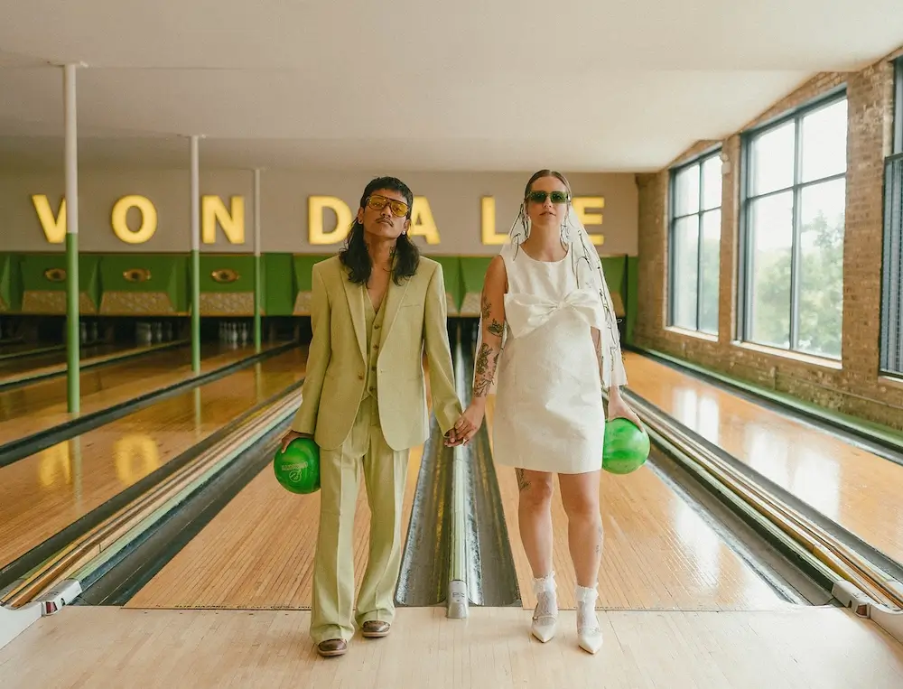 Couple in modern wedding attire holding green bowling balls on a vintage bowling alley lane with mid-century signage in the background.