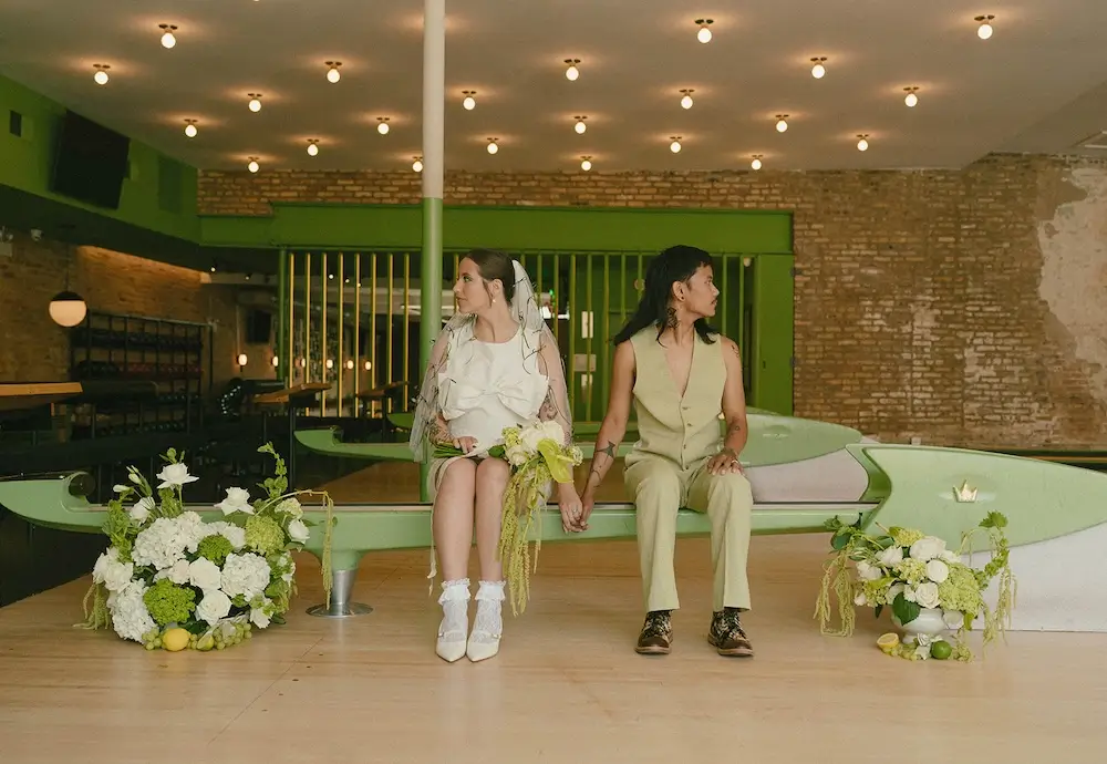 Couple seated on a vintage green bench at a mid-century bowling alley wedding, surrounded by lush green-and-white floral arrangements and retro brick interiors.